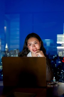 Young businesswoman working late on a laptop inside a modern manhattan office, illuminated by the