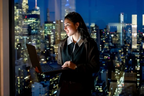 Young businesswoman stands by a floor to ceiling window at night, holding a laptop and smiling at