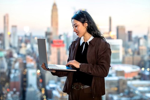 Young businesswoman working on a laptop, standing with a new york city skyline as a blurred