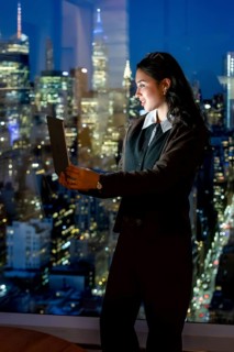 Young businesswoman standing by a high rise window, actively working on a laptop, with the