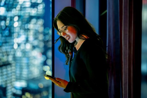 Young businesswoman standing by a skyscraper window at night, focusing on her mobile phone screen