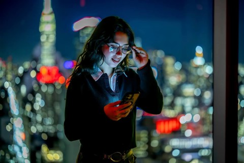 Young executive woman checking her mobile phone, adjusting her eyeglasses by a large window with a