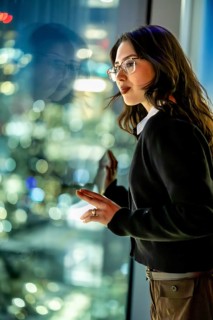 Young businesswoman standing by a skyscraper window, looking thoughtfully at the vibrant night city