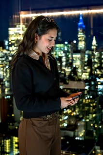Young businesswoman smiling, looking at her smartphone while standing by a window with a stunning
