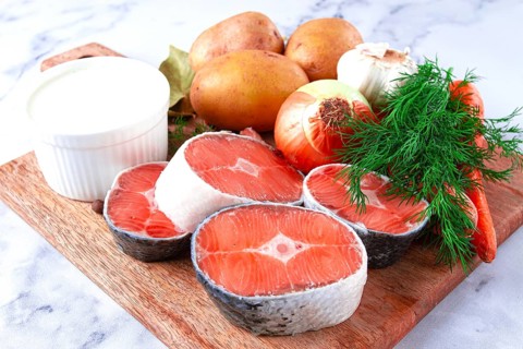 Potatoes, salmon, garlic, onion, and herbs are placed on a wooden cutting board. These ingredients