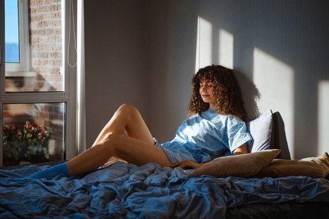 Young woman sits on a bed in a room with sunlight streaming in from a window. She has curly hair
