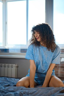 A young woman is sitting on a bed in a simple room. Bright daylight comes through the window,