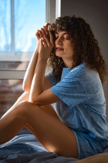 Dreamy woman with curly hair sits on a bed, resting hands together in front of their face. The soft