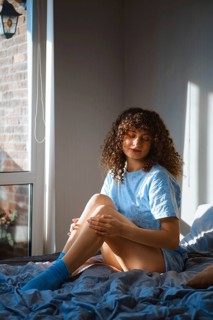 Dreamy woman is sitting on a bed near a window. She is looking down while touching her leg softly.