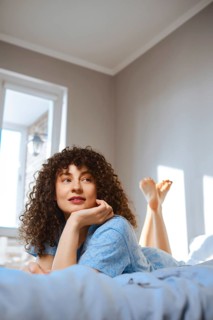 A woman is lying on her stomach on a bed with her legs in the air. She looks thoughtful while