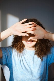 A woman stands in a well-lit room, smiling and covering her face with both hands to block sunlight.