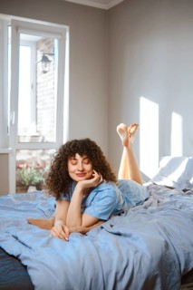 A young woman is lying on her stomach on a bed covered with blue sheet. She rests her chin on her