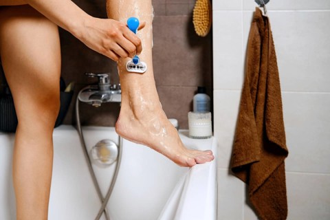 Unrecognizable woman is standing in a bathtub with one leg raised while using a razor to shave.
