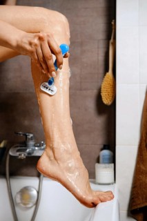 Unrecognizable woman put her leg on the edge of a bathtub and using a razor to shave her leg.