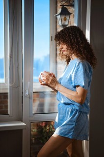 A woman in blue pajamas stands by a window, holding a cup. She looks outside, with sunlight coming