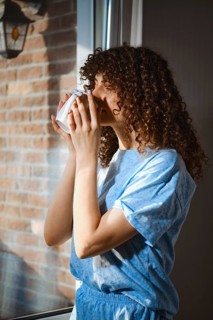 A woman stands by a window and holds a warm drink close to her face. Sunlight shines through the