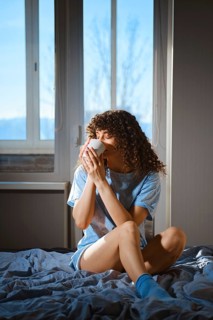 A woman sits on a bed with a blue blanket, holding a white cup. Bright winter sunlight comes