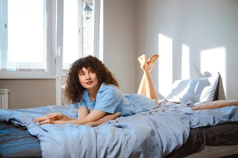 A woman with curly hair lays on a bed with light blue sheets. She looks to the ceiling while