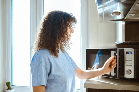 A woman stands by a microwave in a kitchen. She is preparing to heat food. Sunlight enters through