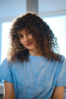 A woman with curly hair sits by a window. She wears a blue shirt and looks down with a thoughtful