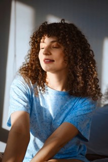 A woman with curly hair sits in a well-lit room. Sunlight streams through the window, creating warm