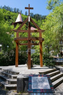 Wooden cross on a pedestal surrounded by green trees and a building, memorial to the tragedy of