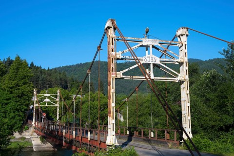 Old suspension bridge stretches across a river surrounded by green landscape, suspension bridge