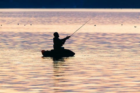 Angler sport fisherman in his belly boat in orange-red lake at sunset, silhouette in calm water,