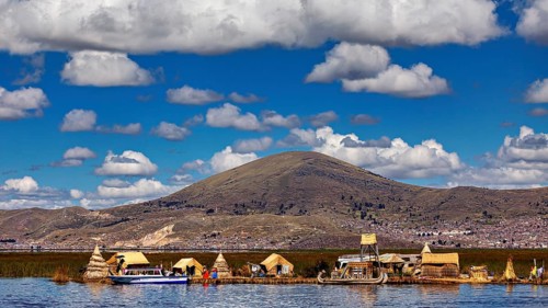 Reed village on the shore of a lake with big mountains in the background under a cloudy sky, The