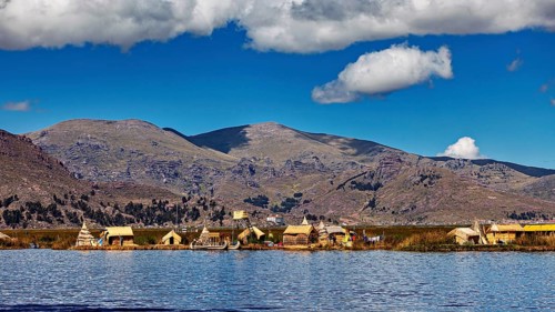 Row of reed huts on a calm lake under a partly cloudy sky and mountain backdrop, The floating reed