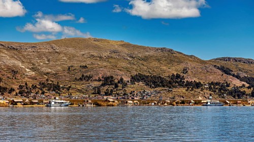 A calm lake in front of a mountainous landscape with boats and a village on the shore under a blue