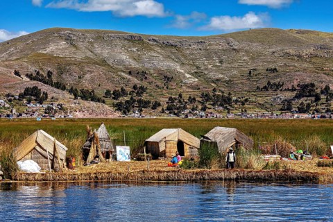 Huts on a reed island in front of a village and mountains, reflecting in the water, The floating