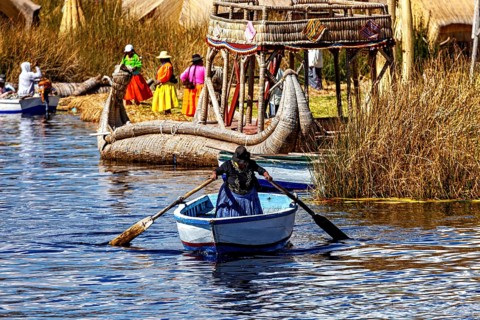 A woman rows in a waterway between large reed canoes and village life, The floating reed islands of