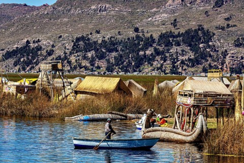 A boat sails on a lake against a backdrop of reed huts and mountains, the floating reed islands of