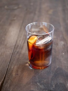Glass of dark alcoholic cocktail with ice and orange zest on a rustic wooden table
