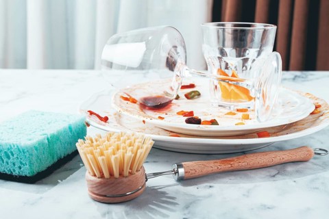 Dishes are stacked on a kitchen counter with food remnants after a meal. A cleaning brush and