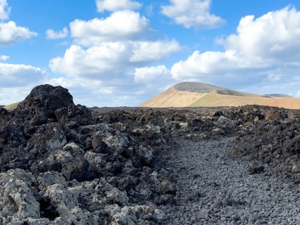Hiking trail through lava rock to Caldera Blanca volcano against blue sky with white clouds, Mancha