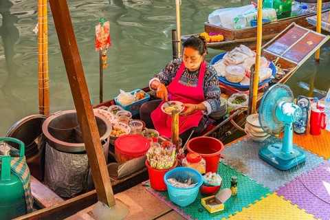 Elderly woman wearing mask offers food on her wooden boat, Damneon Saduak floating markets channel,