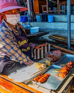 Elderly woman wearing mask sells barbecue skewers on her wooden boat on the Damneon Saduak floating
