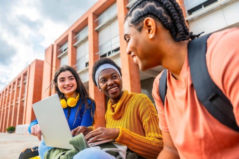 Diverse group of university students collaborating and smiling around a laptop outdoors on campus,