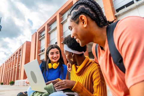 Three diverse students are working together on a laptop computer outside a university building,