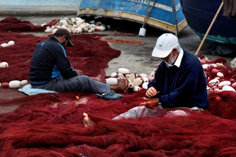 Fishermen repair nets in harbor surrounded by blue boats and tools, Essaouira, Marrakesh-Safi,