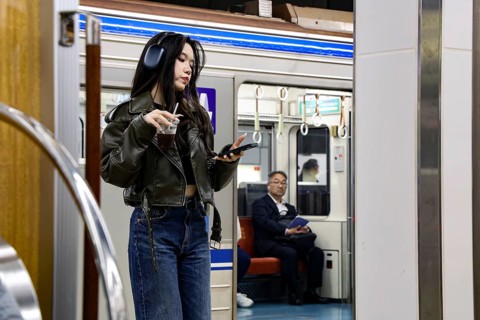 Woman standing in Osaka subway looking at her smartphone while a man sits in the background, Osaka,