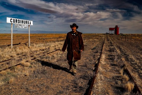 Man wearing cowboy clothes on abandoned tracks in front of station sign and water tower, Oodnadatta