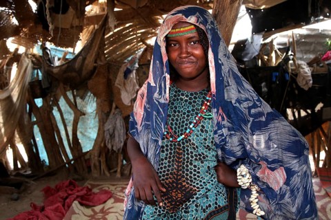 Aishe stands in Muhammad's hut wearing traditional clothes surrounded by coloured fabric in