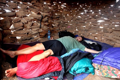 Person lying in a sleeping bag in a simple stone hut, zero