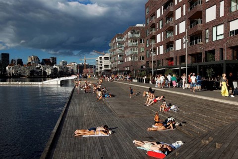 People relaxing on wooden walkway in the sun surrounded by new buildings, Oslo, Norway