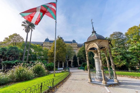 Basque ikurrina flag waving over historic gipuzkoa square in san sebastian, framed by lush gardens,