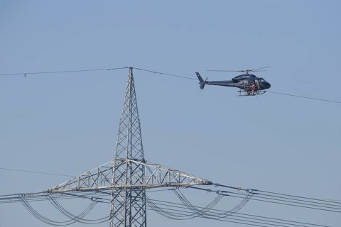 A helicopter carries out technical work on power lines next to a mast under a blue sky, an employee
