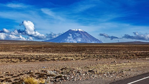 Volcano in a wide desert landscape under blue sky with clouds, snow-capped summit, The Andean